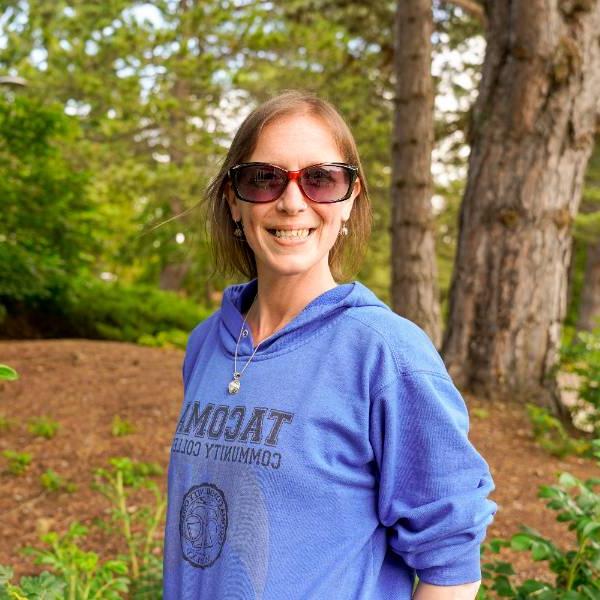 A headshot of Leigh. 她在树林的背景下微笑, 戴着长方形太阳镜，穿着一件宝蓝色的“澳门威尼斯人在线赌场”运动衫.